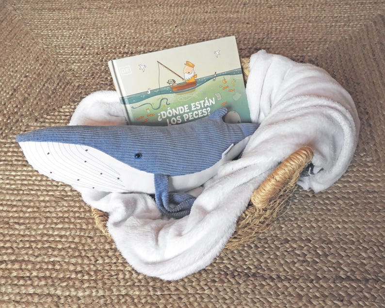 Peut inclure: Un panier en osier contient une peluche baleine bleue, un livre pour enfants et une couverture blanche. La couverture du livre pr&eacute;sente une illustration d'une personne p&ecirc;chant depuis un bateau. Le texte du livre est "&iquest;D&oacute;nde est&aacute;n los peces?"