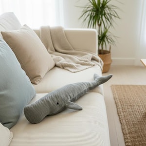 K&ouml;nnte beinhalten: Ein graues Wal-Kissen aus Cord liegt auf einem wei&szlig;en Sofa, zusammen mit hellblauen und beigen Kissen. Eine beige Decke ist &uuml;ber das Sofa drapiert. Ein Holztisch und ein gewebter Teppich befinden sich im Hintergrund.