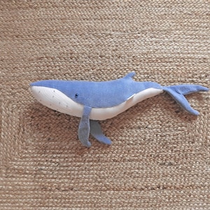 Peut inclure: Une peluche baleine bleue et blanche avec un corps textur&eacute; en forme de velours c&ocirc;tel&eacute;. Le jouet a un ventre blanc et un petit &oelig;il bleu fonc&eacute;. Il est pos&eacute; sur un tapis beige tiss&eacute;.