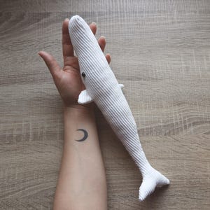Puede incluir: Un juguete de peluche de ballena blanca con un ojo negro, sostenido en la mano de una persona. La persona tiene un tatuaje de media luna en la muñeca.