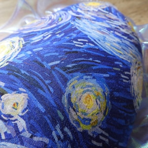 Puede incluir: Primer plano de un art&iacute;culo de tela con un estampado de "La noche estrellada" de Van Gogh. El dise&ntilde;o incluye pinceladas azules, amarillas y blancas arremolinadas. El art&iacute;culo est&aacute; parcialmente cubierto por un material transl&uacute;cido e iridiscente.