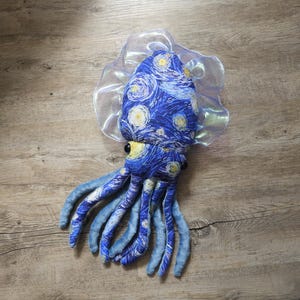 Könnte beinhalten: Ein Plüsch-Tintenfisch-Spielzeug mit einem blau-gelben Sternennacht-Design und einem durchscheinenden, gerüschten Kragen. Der Tintenfisch hat blaue Tentakel und gelbe Augen. Das Spielzeug liegt auf einer Holzoberfläche.