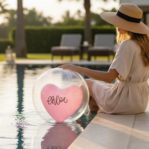 May include: A transparent beach ball featuring a pink heart-shaped insert with the name "Chloe" written on it. The ball is near a swimming pool. A person wearing a hat and dress is sitting by the pool.
