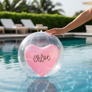 May include: A clear, spherical inflatable beach ball containing a pink, heart-shaped balloon inside. The pink heart features the name Chloe written in black cursive script. The ball is floating on the surface of a blue swimming pool.