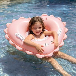 May include: A pink heart-shaped inflatable pool ring with a scalloped edge. The ring has white handles and the name "Evie" written on it. A young girl is sitting inside the ring in a swimming pool.