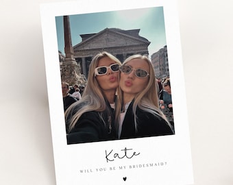 May include: A white invitation card with a photo of two women wearing sunglasses, posing for a selfie. The text "Kate, Will you be my bridesmaid?" is written below the photo, with a heart symbol.