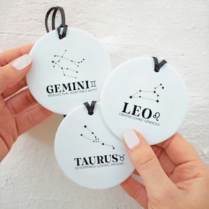 May include: Three white ceramic ornaments featuring black zodiac signs and descriptions. The signs are Gemini, Leo, and Taurus.