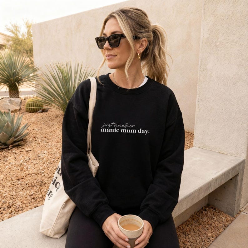 May include: Black sweatshirt with the text "just another manic mum day." The sweatshirt is worn with black sunglasses and a white tote bag. The person is holding a cup of coffee.