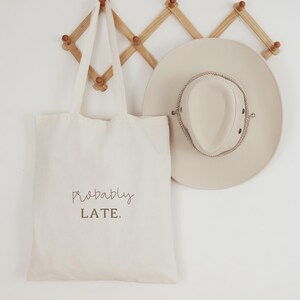 May include: A cream canvas tote bag with the words "Probably LATE" printed on it, hangs beside a beige hat with a rope trim. The hat has a wide brim and is made of a light-colored material. The bag has long handles.