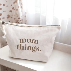 May include: Cream canvas cosmetic bag with a zip closure. The bag has the words "mum things." printed in brown, serif font. The bag is on a white surface.