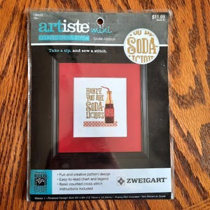 May include: A cross-stitch kit featuring a framed design with the text "Honey, You Are Soda-licious" and an illustration of a soda bottle. The kit includes instructions and is designed for counted cross-stitch. Finished design size is 5in x 6in.