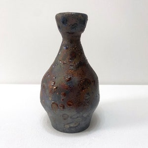 Puede incluir: Un jarrón de cerámica marrón oscuro y gris con una superficie texturizada. El jarrón tiene un cuerpo redondo y un cuello estrecho.