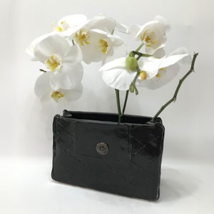 以下が含まれることがあります： キルティングのデザインと装飾的なボタンが付いた黒いセラミックの花瓶。白い蘭が花瓶にアレンジされています。