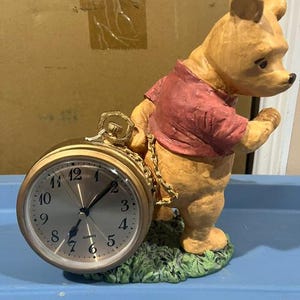 May include: A decorative figurine of a yellow bear wearing a red shirt, standing on a green grassy base. The bear holds a gold-toned round quartz clock with a metallic face, black numerals, and black hands, connected by a gold-colored chain.