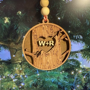 Op de afbeelding: Een houten kerstversiering in de vorm van een boom met een hart in het midden. Het hart heeft de initialen "W+R" erin gegraveerd.