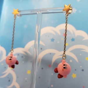 Może przedstawiać: Para różowych i złotych kolczyków wiszących z postacią z kreskówki i zawieszką w kształcie gwiazdki.