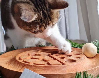 May include: A tabby cat with white paws plays with a wooden cat toy. The toy is a circular wooden board with a maze carved into it. A wooden ball is on the board. The toy is labeled "Waldreif" with a mountain logo.