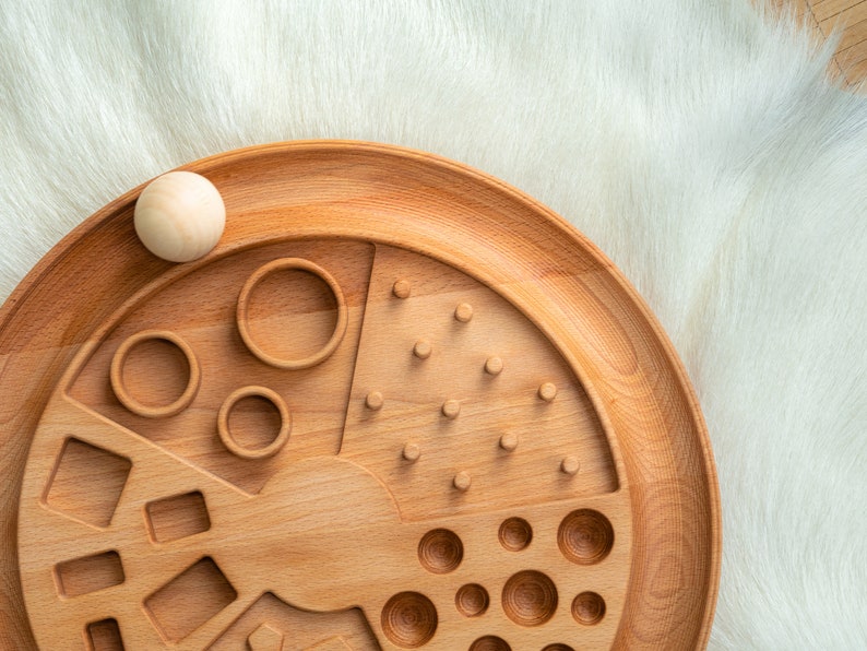 May include: A wooden toy with different shapes and patterns for children to explore. The toy has a circular base with a variety of cutouts, including circles, squares, and triangles. There are also pegs and holes for sorting and matching.