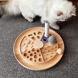 May include: A wooden enrichment board for cats with a variety of textures and shapes. The board has a round shape and is made of light wood. It features a variety of activities for cats, including a maze, a puzzle, and a treat dispenser. The board also includes a small, gray felt ball and a white cardboard tube.