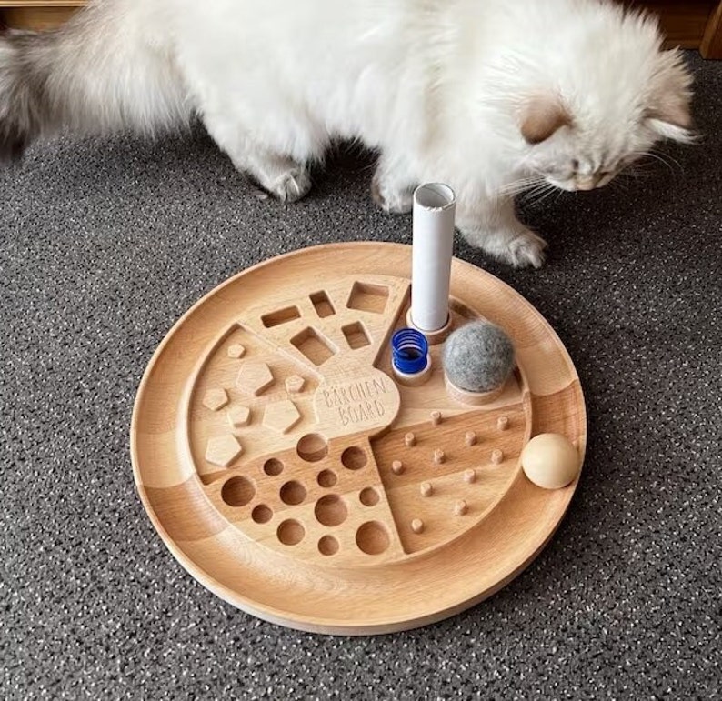 May include: A wooden enrichment board for cats with a variety of textures and shapes. The board has a round shape and is made of light wood. It features a variety of activities for cats, including a maze, a puzzle, and a treat dispenser. The board also includes a small, gray felt ball and a white cardboard tube.
