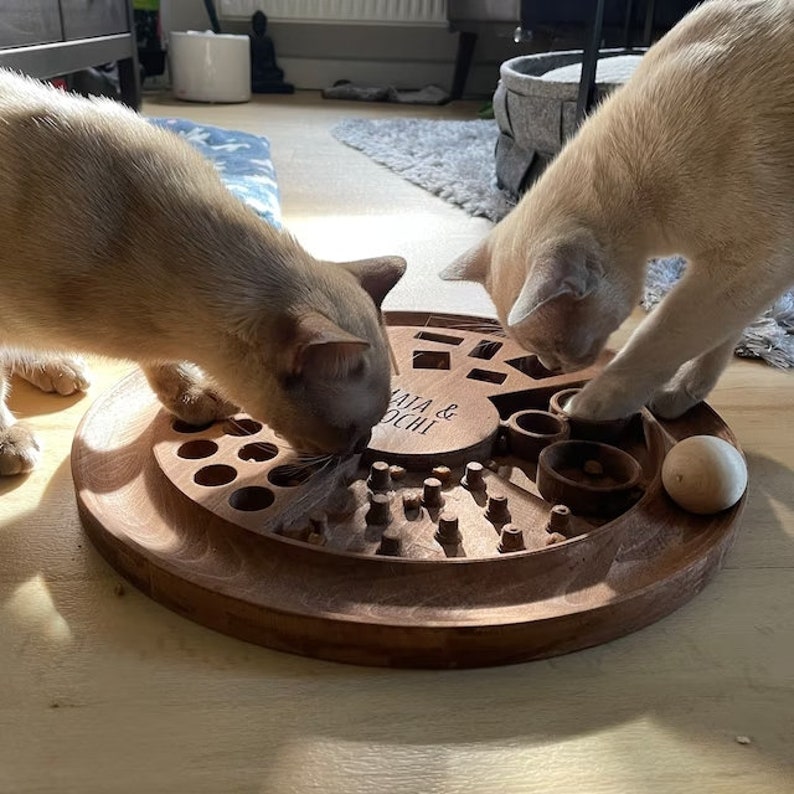 May include: Two brown cats playing with a wooden cat toy with a variety of compartments and a ball. The toy is engraved with the text "Maya & Mochi".