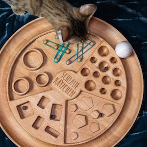 May include: A wooden cat toy with different shapes and textures. The toy has a white ball, a section with small holes, a section with different shapes, and a section with a maze. The toy is labeled "Schaue K&auml;tzchen".