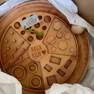 May include: A wooden tray with different shaped cutouts, including circles, squares, and hexagons. The tray is labeled "NASC KATZE" and has a small heart symbol. The tray is made by Waldreif.