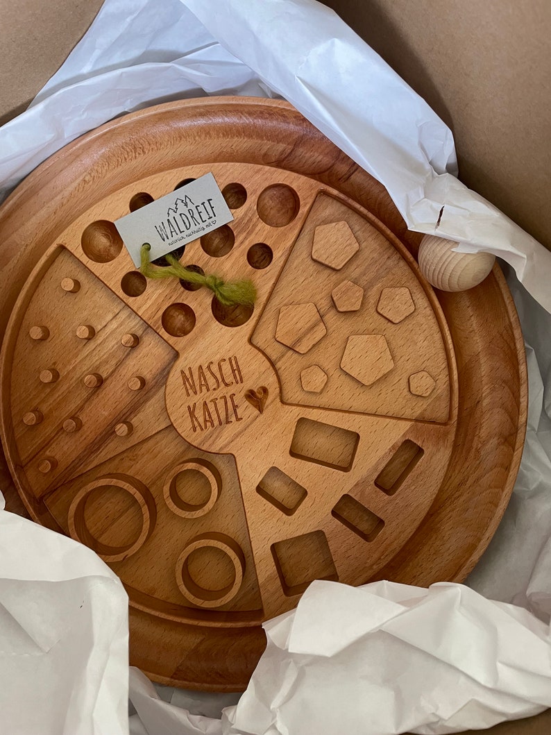 May include: A wooden tray with different shaped cutouts, including circles, squares, and hexagons. The tray is labeled "NASC KATZE" and has a small heart symbol. The tray is made by Waldreif.