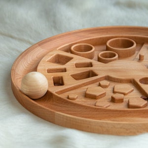 May include: A wooden toy with various shapes and sizes cut out of the surface. The toy is designed for children to learn about shapes and sizes. A wooden ball is sitting next to the toy.