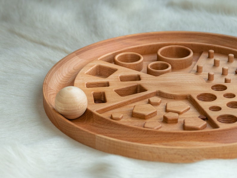May include: A wooden toy with various shapes and sizes cut out of the surface. The toy is designed for children to learn about shapes and sizes. A wooden ball is sitting next to the toy.