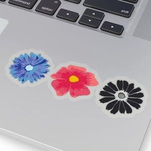 May include: Three floral stickers on a laptop surface. The stickers feature a blue flower, a red flower with a yellow centre, and a black and white flower. The laptop keyboard is visible in the background.