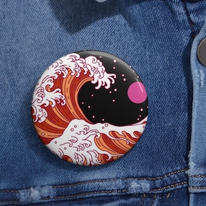 May include: A round button pin featuring a stylized wave design in shades of orange, red, and white, set against a black background with a pink circle and white stars. The pin is displayed on a denim jacket.