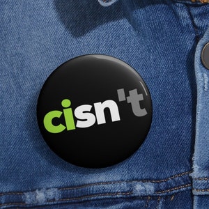 May include: A black button with the text "cisn't" in white and green letters. The button is pinned to a blue denim jacket.