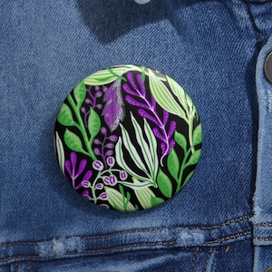 May include: A round button with a black background and a colorful floral pattern. The pattern features green, purple, and white leaves and flowers.