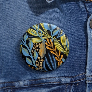May include: A round button with a black background and a colorful floral pattern. The pattern features yellow, blue, and orange leaves and stems.