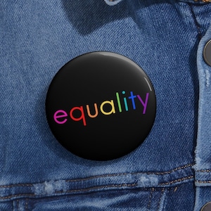 May include: A black button pin with the word "equality" in rainbow colors. The pin is set against a denim jacket.