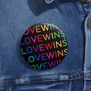 Puede incluir: Un botón redondo y negro con las palabras "LOVE WINS" en colores del arcoíris. El botón está sobre una chaqueta vaquera azul.