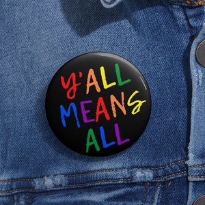 Peut inclure: Un bouton de badge noir avec l'inscription "Y'ALL MEANS ALL" en couleurs arc-en-ciel. L'épingle est ronde et est posée sur une veste en jean.