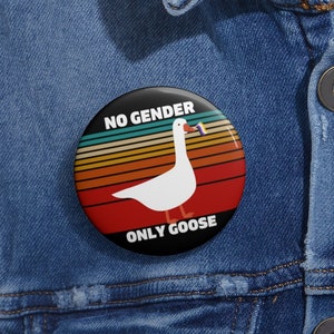 May include: A round button with a retro design featuring a white goose holding a pride flag in its beak. The button has the text "NO GENDER ONLY GOOSE" in black text on a black background.