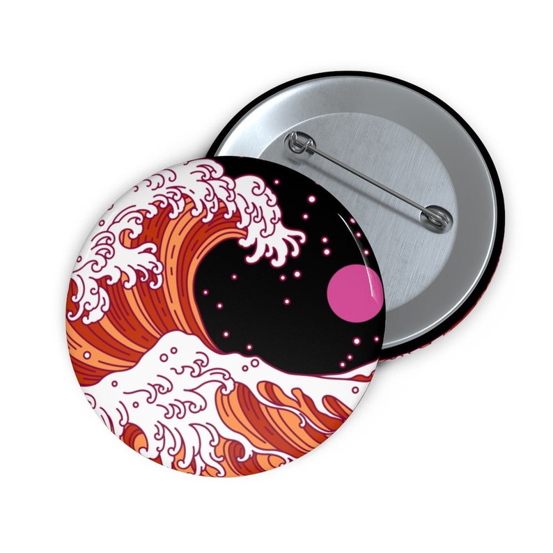 May include: A button pin with a design featuring a stylized orange and white wave against a black background with a pink circle and white dots. The pin has a silver back and a safety pin.