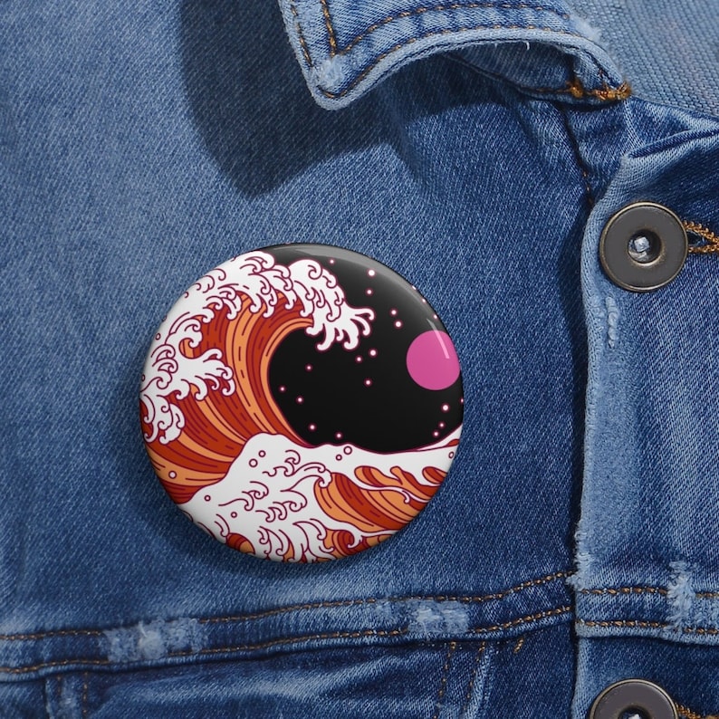 May include: A round button pin featuring a stylized wave design in shades of orange, red, and white, set against a black background with a pink circle and white stars. The pin is displayed on a denim jacket.