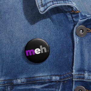 May include: A black button with the word "meh" in white and purple letters. The button is pinned to a blue denim jacket.