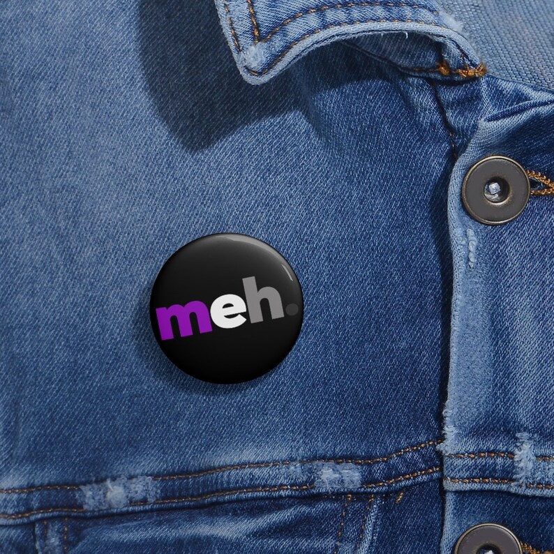 May include: A black button with the word "meh" in white and purple letters. The button is pinned to a blue denim jacket.