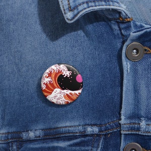 May include: A round button pin featuring a stylized wave design in shades of red, orange, and white, with a black background and a pink circle. The pin is set against a denim jacket.