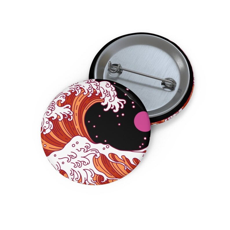 May include: Two round pinback buttons. One button features a stylized wave design in shades of orange and white against a black background with a pink circle. The other button is silver with a pin back.
