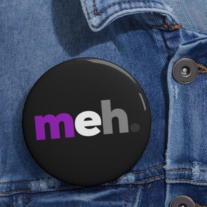 May include: A black button with the word "meh" in purple, white, and gray letters. The button is on a denim jacket.