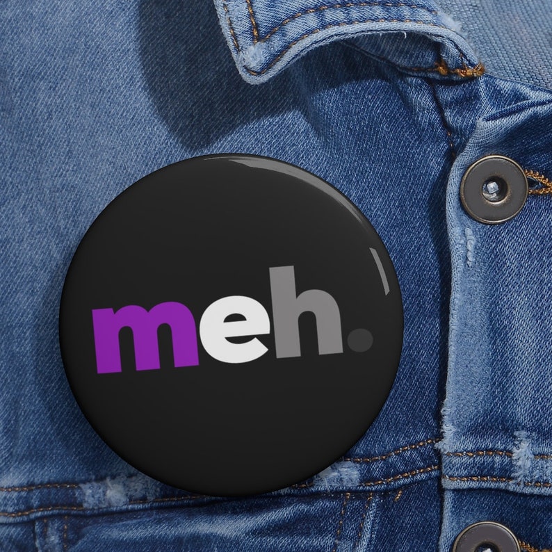 May include: A black button with the word "meh" in purple, white, and gray letters. The button is on a denim jacket.