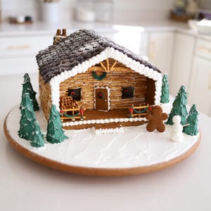 Op de afbeelding: Een gingerbreadhuisje versierd als een blokhut met een bruin dak, witte glazuurranden en groene bomen. Een gingerbreadman staat voor het huis.