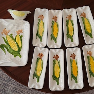 May include: A ceramic dinnerware set featuring a large rectangular serving platter, a small gravy boat, and eight long, narrow corn-shaped dishes. Each piece is white with a textured surface and a raised, hand-painted yellow corn cob design.