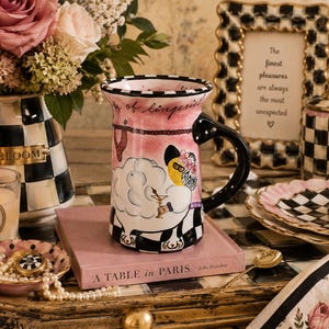 Puede incluir: Una taza de cerámica con una ilustración caprichosa de una oveja en una bañera sobre un fondo rosa, con una base de cuadros blancos y negros y un asa negra. La taza muestra el texto "of lingerie" y se apoya sobre un libro rosa A TABLE IN PARIS.